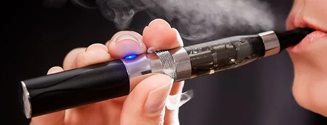 Primer plano de una persona con un vapeador negro y metálico en los labios, con luz azul encendida y vapor alrededor.