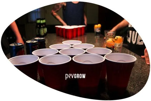 Vasos rojos dispuestos en triángulo sobre una mesa, con otro grupo al fondo, latas, bebidas y varias personas alrededor.