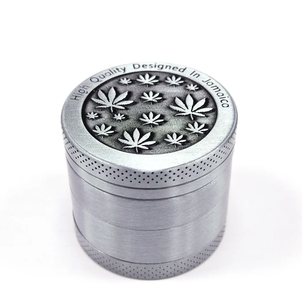 Grinder Metal Leaves plateado y cilíndrico, con hojas de cannabis en relieve sobre la tapa oscura y bordes de agarre punteados.