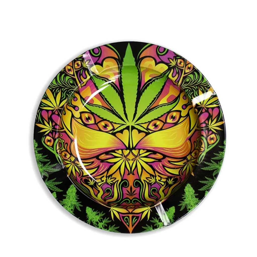 Cenicero redondo Basic Leaf visto desde arriba, con una hoja de cannabis verde y motivos simétricos amarillos y rosas sobre fondo negro.