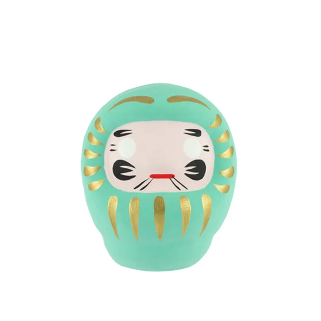 Daruma turquesa de frente, con adornos dorados, rostro claro, ojos sin pupilas y detalles negros y rojos. Daruma turquesa de frente, con adornos dorados, rostro claro, ojos sin pupilas y detalles negros y rojos.