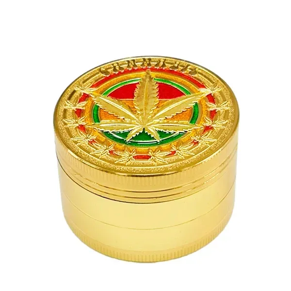 Grinder Rasta Gold dorado con hoja de cannabis en relieve sobre franjas rojas, amarillas y verdes, visto en perspectiva.