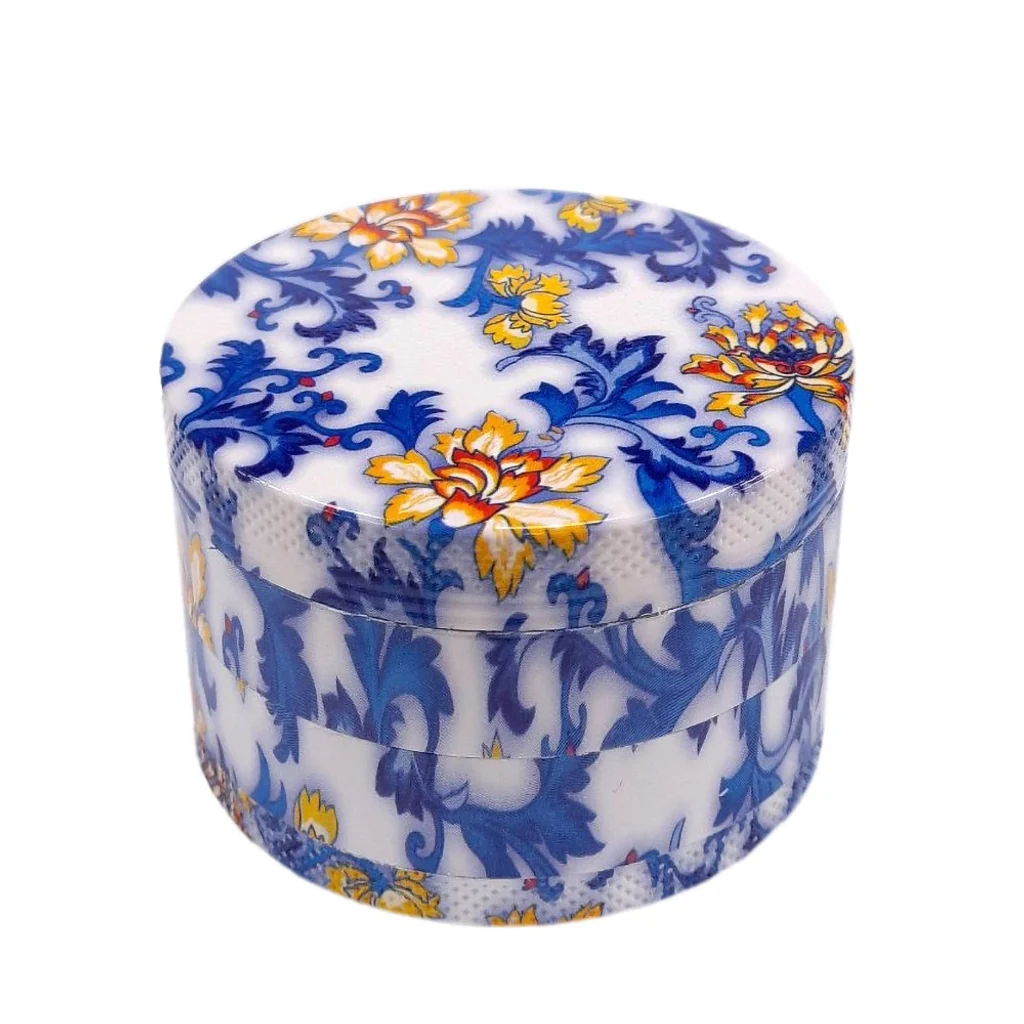 Grinder Flowers cerrado y cilíndrico, visto en perspectiva, con fondo blanco, motivos vegetales azules y flores amarillas y naranjas.