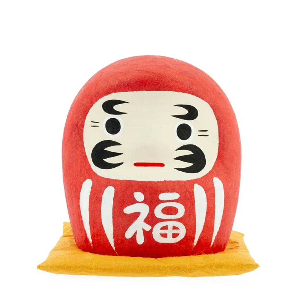 Daruma rojo de cuerpo redondeado sobre cojín amarillo, con rostro crema, detalles negros y carácter blanco en el frontal. Daruma rojo de cuerpo redondeado sobre cojín amarillo, con rostro crema, detalles negros y carácter blanco en el frontal.