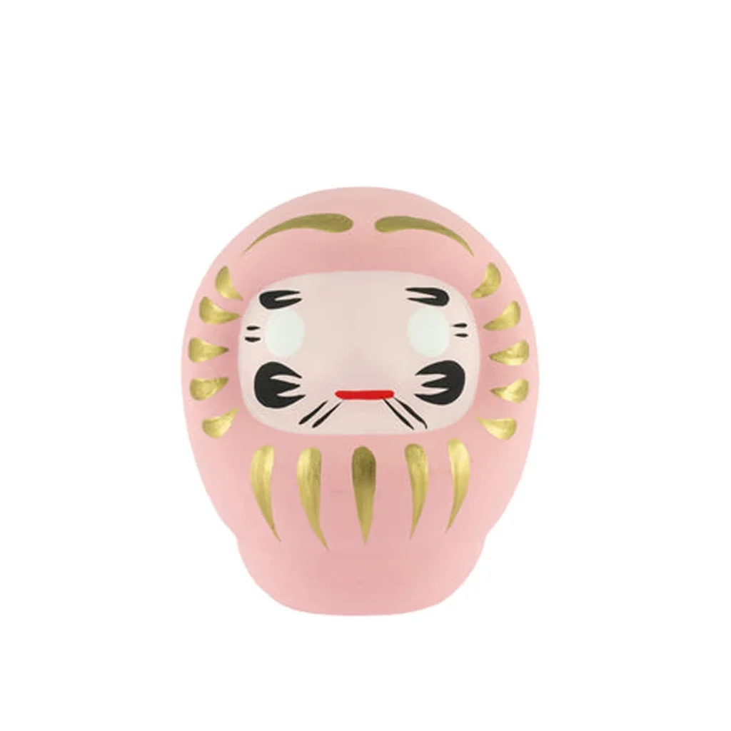 Daruma rosa visto de frente, con detalles dorados, cejas y bigote negros, ojos blancos sin pupilas y una pequeña boca roja. Daruma rosa visto de frente, con detalles dorados, cejas y bigote negros, ojos blancos sin pupilas y una pequeña boca roja.