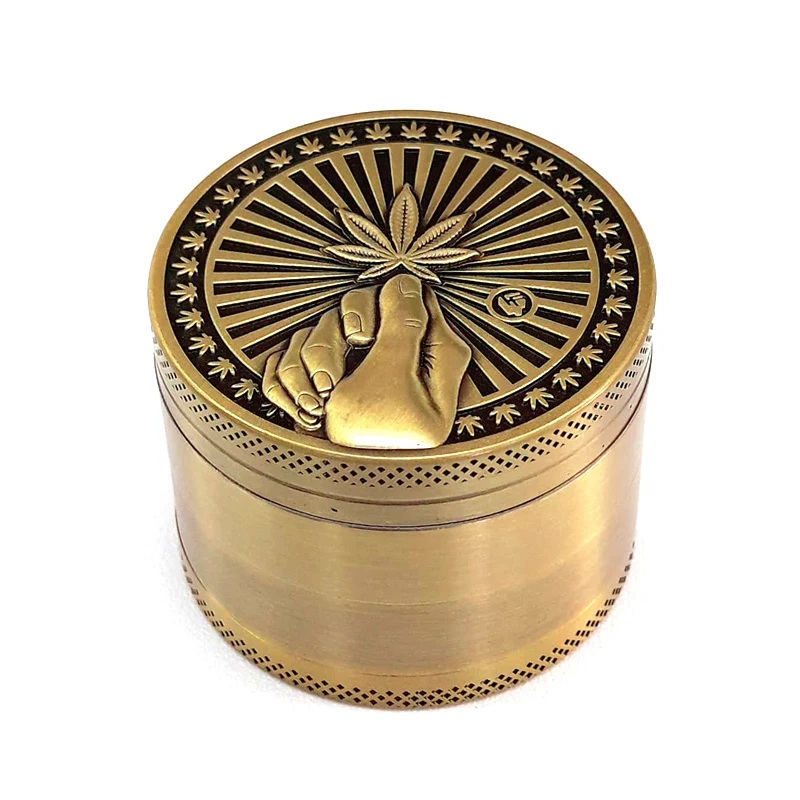 Grinder Gold Hope de acabado dorado, con una mano sosteniendo una hoja de cannabis en relieve y motivos radiales en la tapa.