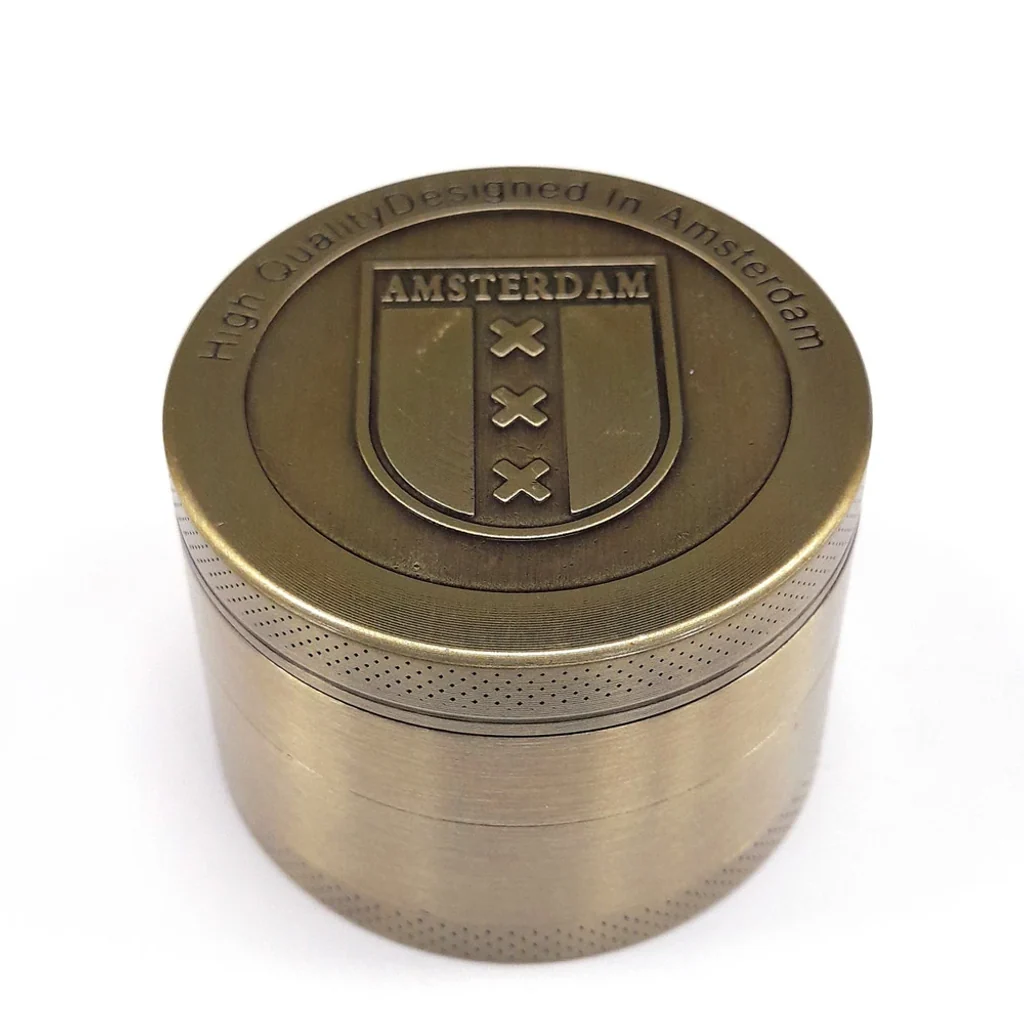 Grinder Gold Shield cerrado, de tono dorado envejecido, con escudo de Amsterdam y cruces en relieve sobre la tapa.