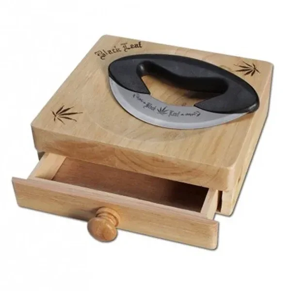 Tabla de madera Black Leaf con cajón abierto, motivos de hojas grabados y cuchilla curva con mango negro apoyada encima.