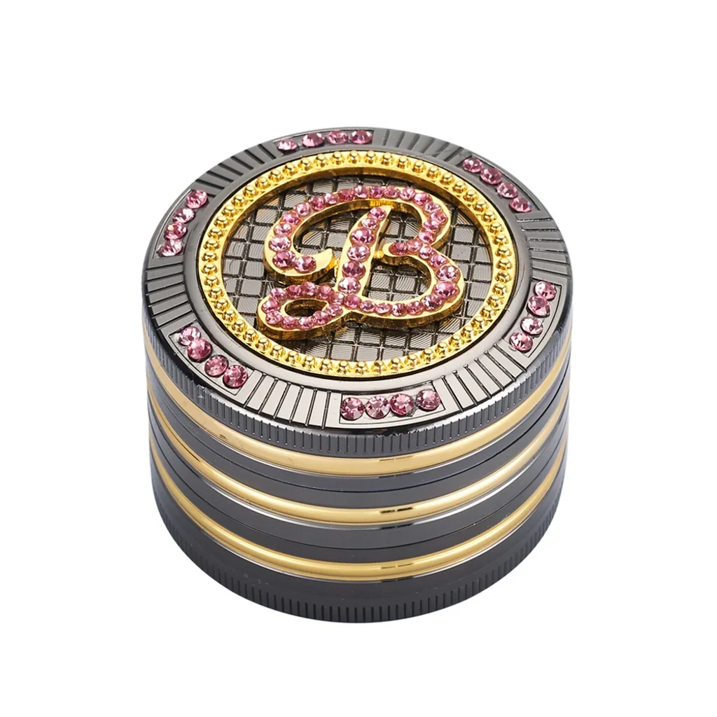 Grinder cilíndrico gris oscuro con bandas doradas y tapa decorada con una letra B y piedras rosas y amarillas, visto en perspectiva.