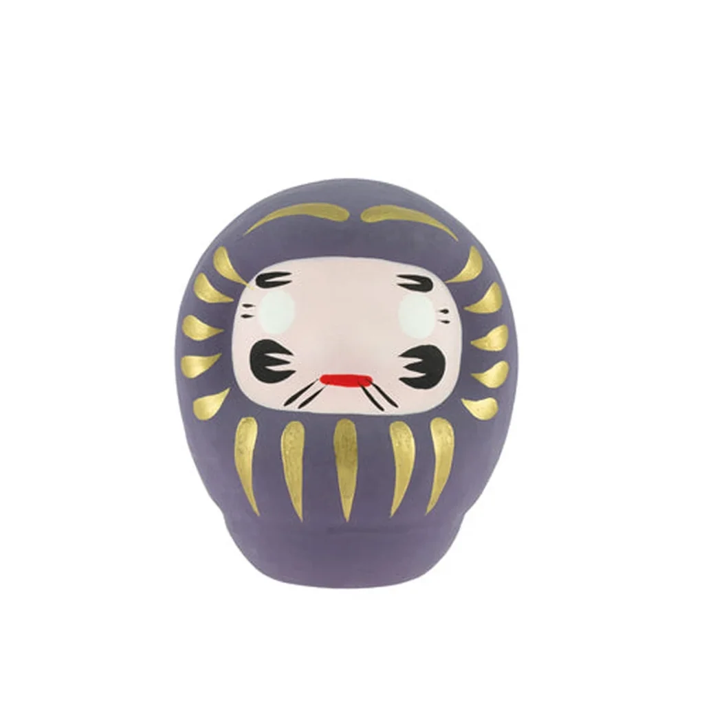 Daruma tradicional violeta de frente, con rostro claro, detalles negros, boca roja y adornos dorados sobre fondo blanco. Daruma tradicional violeta de frente, con rostro claro, detalles negros, boca roja y adornos dorados sobre fondo blanco.