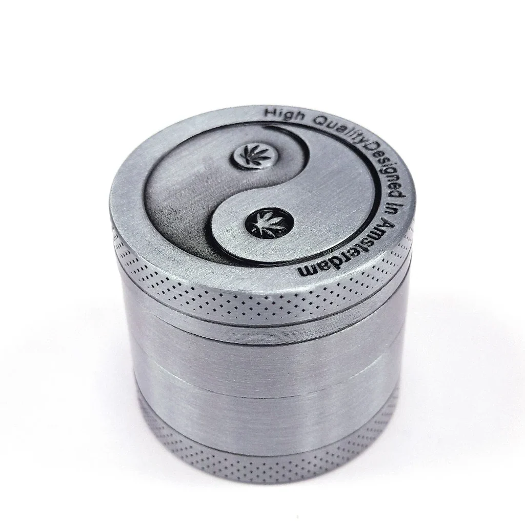 Grinder metálico plateado cerrado, visto en perspectiva, con símbolo yin-yang y dos hojas de cannabis en la tapa.