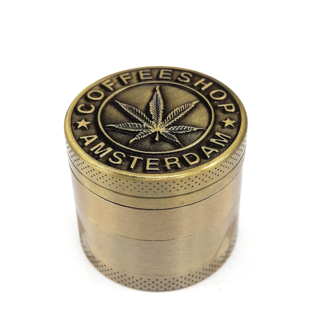 Grinder dorado cilíndrico con bordes punteados y tapa en relieve con una hoja de cannabis y el texto «Coffeeshop Amsterdam».