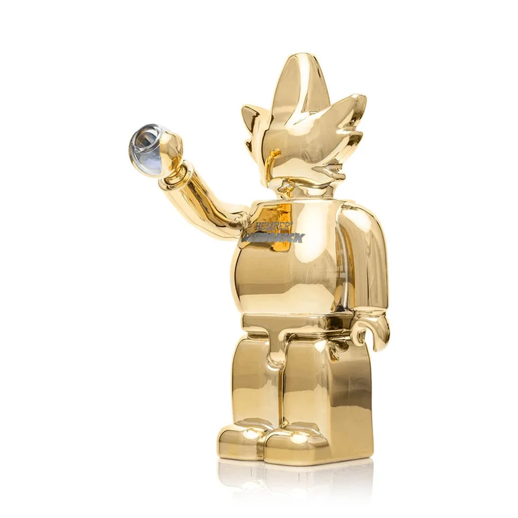 Bong HEMPER Weedbrick XL dorado con forma de figura articulada, cabeza de hoja y un brazo elevado que sostiene una cazoleta.