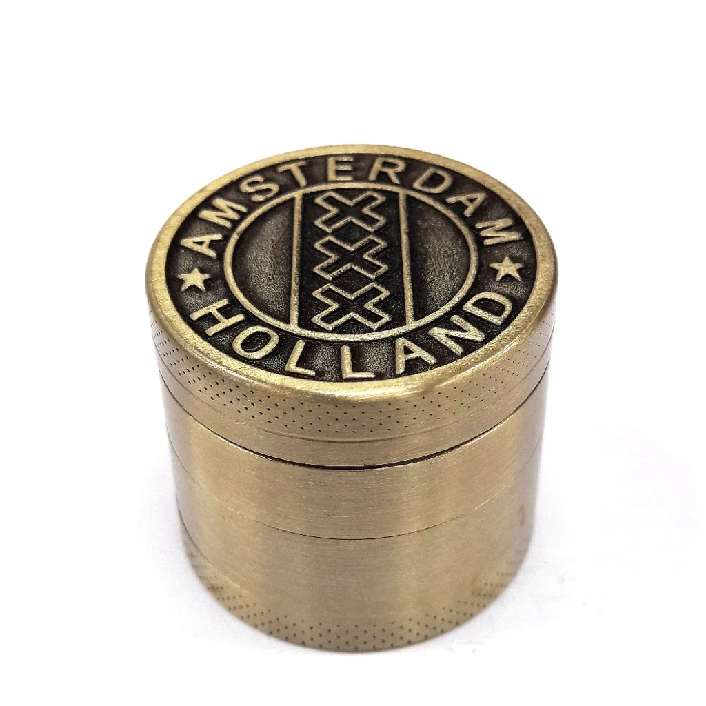 Grinder cilíndrico dorado con varias secciones y tapa grabada con Amsterdam, Holland y tres cruces, visto en perspectiva.
