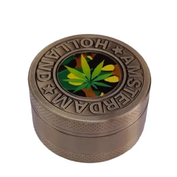 Grinder metálico de tono dorado envejecido, con hoja de cannabis multicolor en la tapa y texto Amsterdam Holland en relieve.