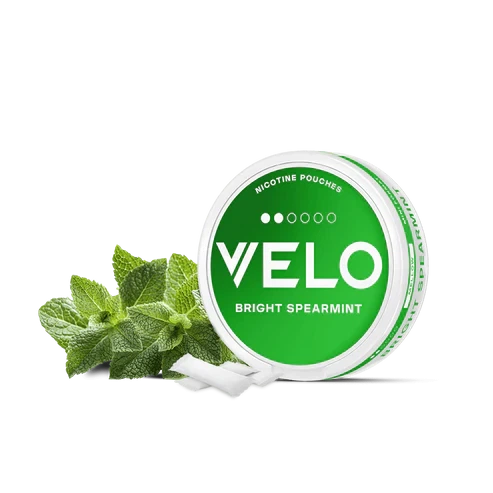 Envase circular verde y blanco de VELO Bright Spearmint, con hojas de menta y bolsitas blancas delante, sobre fondo negro. Envase circular verde y blanco de VELO Bright Spearmint, con hojas de menta y bolsitas blancas delante, sobre fondo negro.