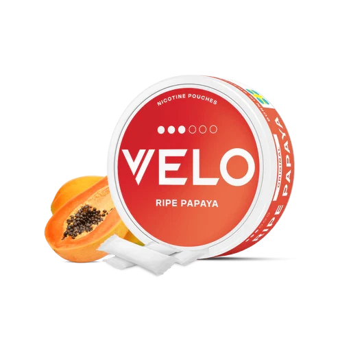 Envase redondo de VELO Ripe Papaya con etiqueta roja y naranja, junto a una papaya abierta y varias bolsas blancas sobre fondo negro. Envase redondo de VELO Ripe Papaya con etiqueta roja y naranja, junto a una papaya abierta y varias bolsas blancas sobre fondo negro.