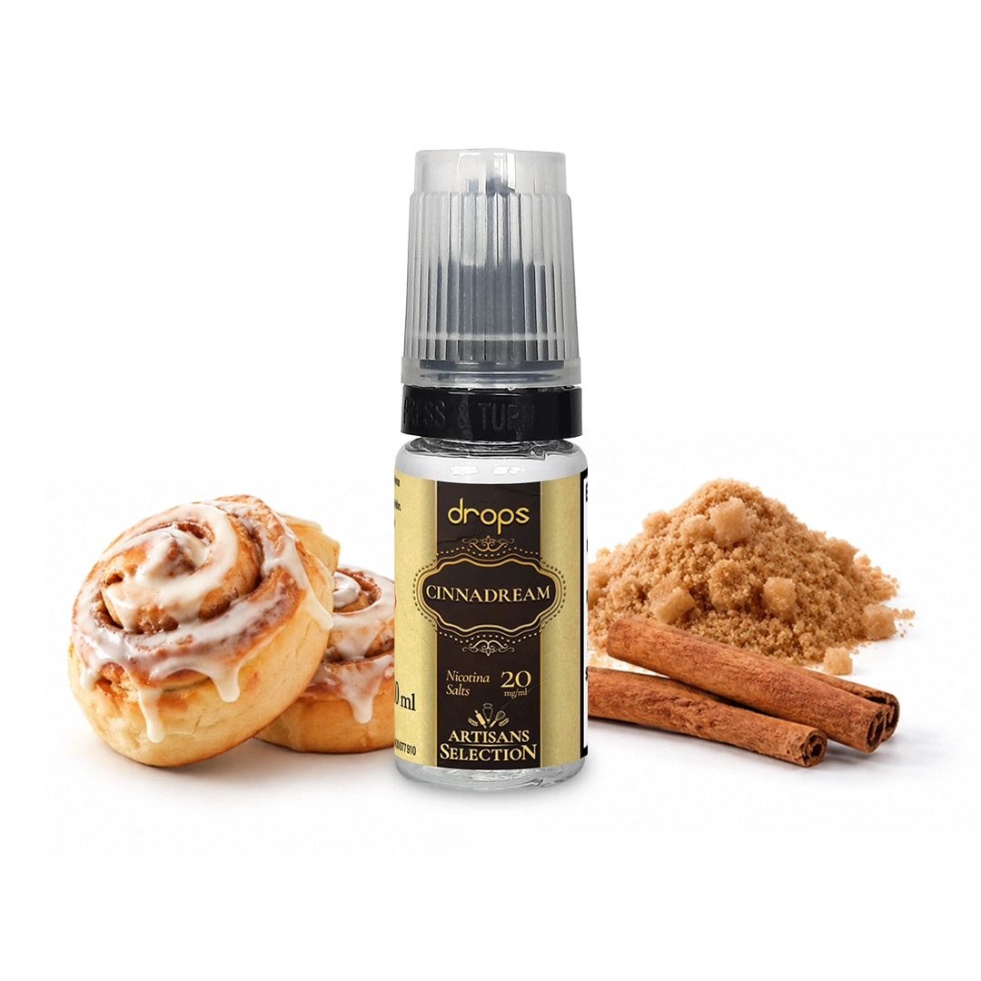 Frasco de Drops Cinnadream con etiqueta dorada y negra, junto a bollos glaseados, canela en rama y un montón de azúcar moreno.