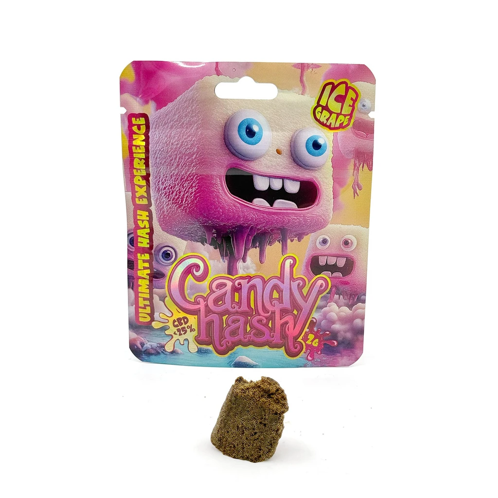 Envase rosa y amarillo de Candy Hash Ice Grape con personaje de ojos azules y un bloque marrón de textura granulada delante.