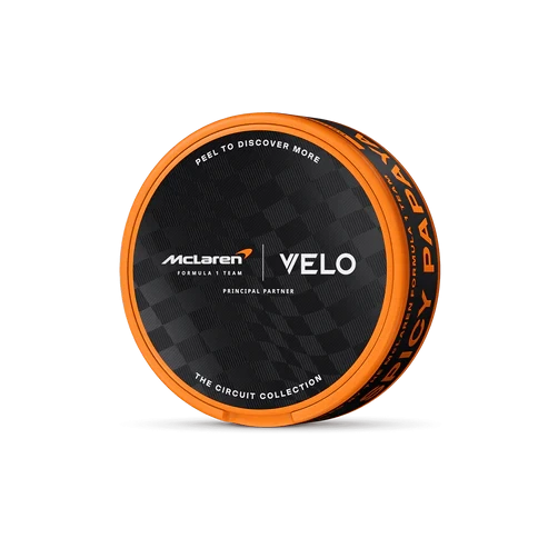 Envase redondo de VELO Spicy Papaya, inclinado sobre fondo negro, con borde naranja y tapa negra con los logotipos de McLaren y VELO. Envase redondo de VELO Spicy Papaya, inclinado sobre fondo negro, con borde naranja y tapa negra con los logotipos de McLaren y VELO.