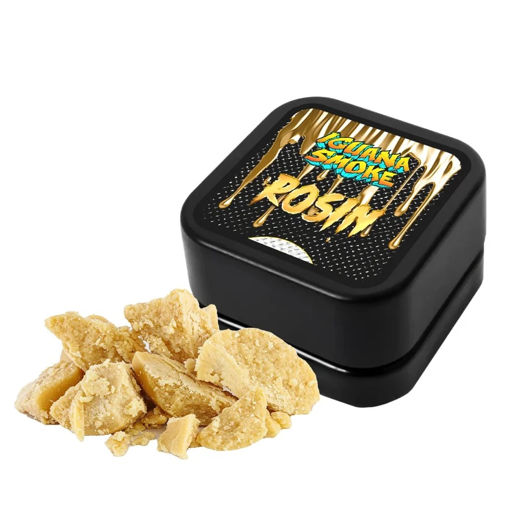 Fragmentos amarillos de aspecto desmenuzable junto a una caja negra de Cold Cure Rosin de Iguana Smoke con etiqueta dorada.