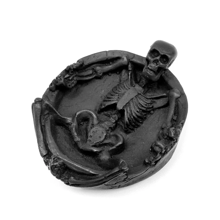 Cenicero negro de forma redondeada con un esqueleto en relieve, cuyo cráneo sobresale del borde, sobre fondo blanco.