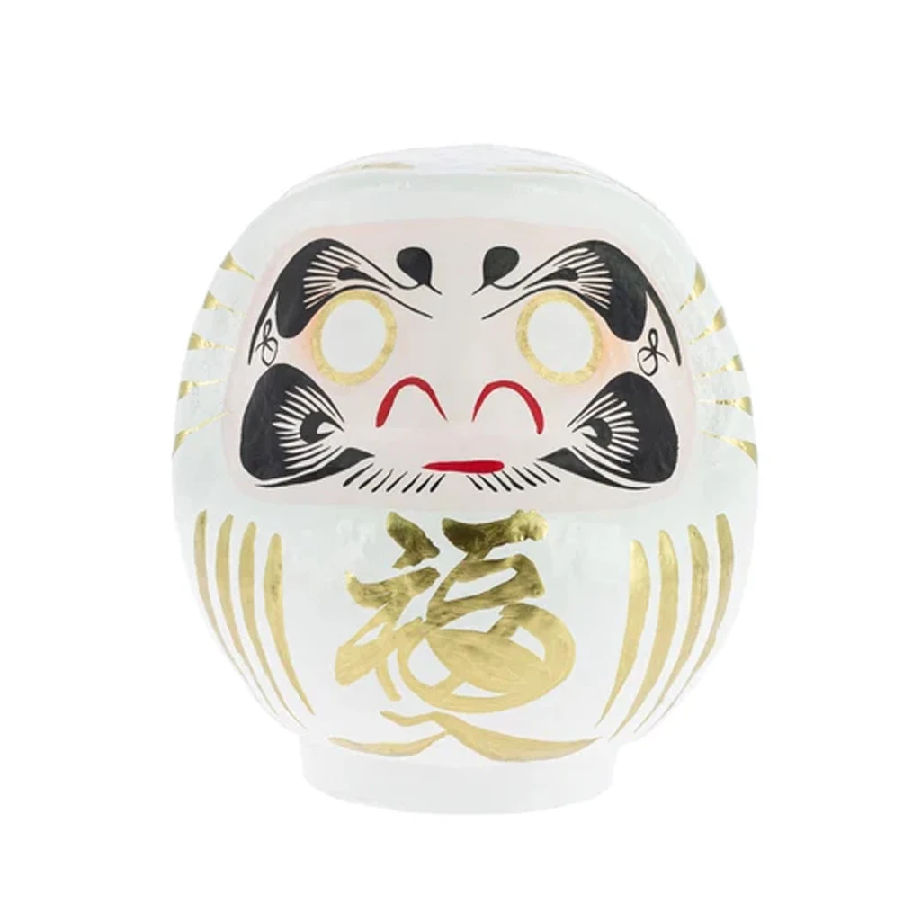 Daruma tradicional blanco visto de frente, con ojos sin pintar, rasgos negros y rojos y un gran carácter dorado en el vientre. Daruma tradicional blanco visto de frente, con ojos sin pintar, rasgos negros y rojos y un gran carácter dorado en el vientre.