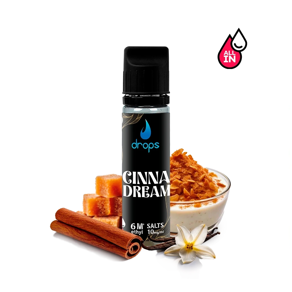 Frasco Drops Cinnadream con etiqueta y tapón negros, junto a ramas de canela, cubos dorados, un cuenco cremoso y una flor blanca.