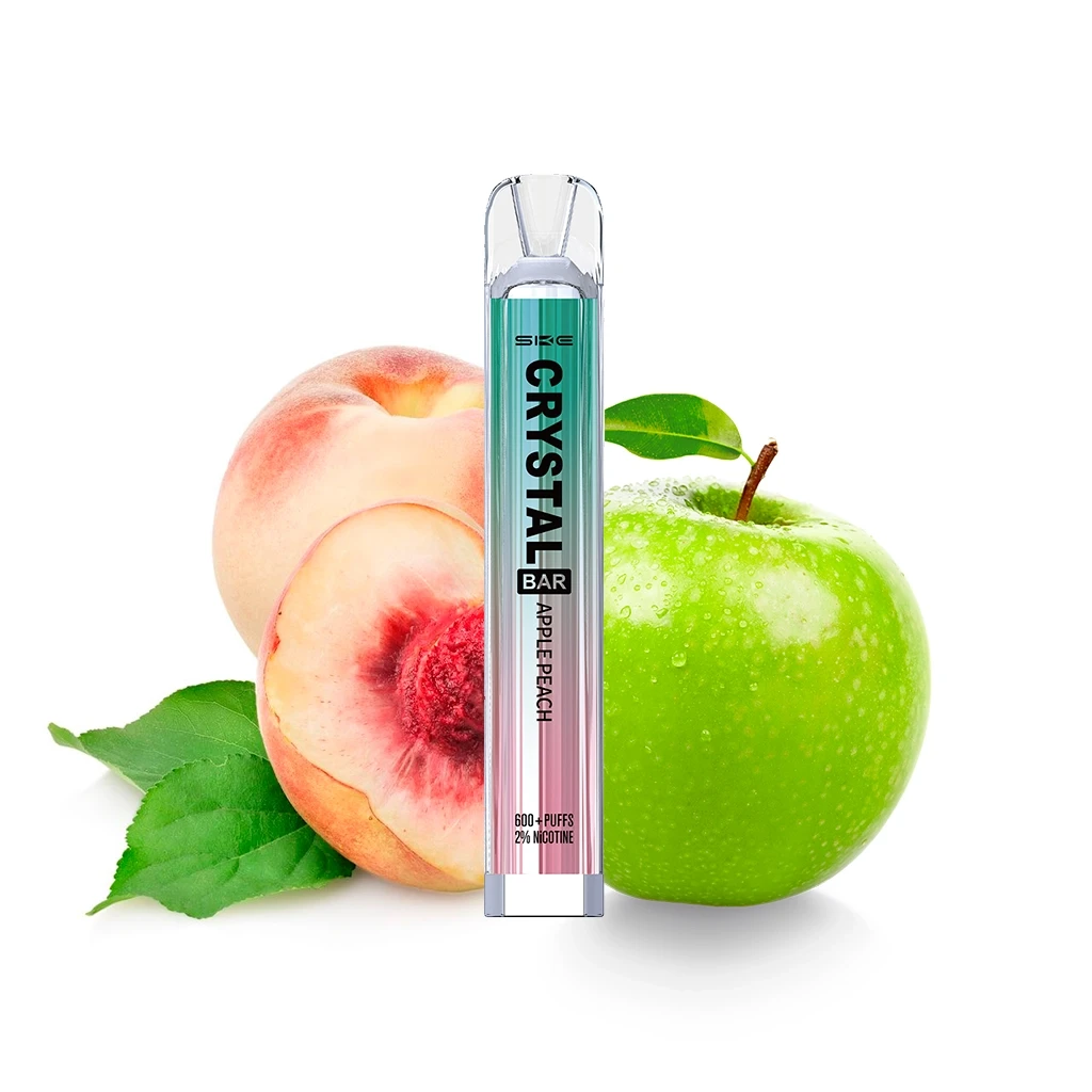 SKE Crystal Bar Apple Peach con cuerpo verde y rosa y boquilla transparente, delante de una manzana verde y melocotones. SKE Crystal Bar Apple Peach con cuerpo verde y rosa y boquilla transparente, delante de una manzana verde y melocotones.
