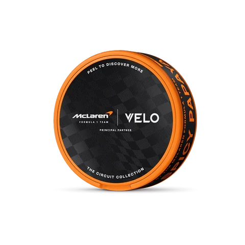 Envase circular VELO Spicy Papaya con borde naranja y tapa negra con los logotipos de McLaren y VELO, sobre fondo negro.
