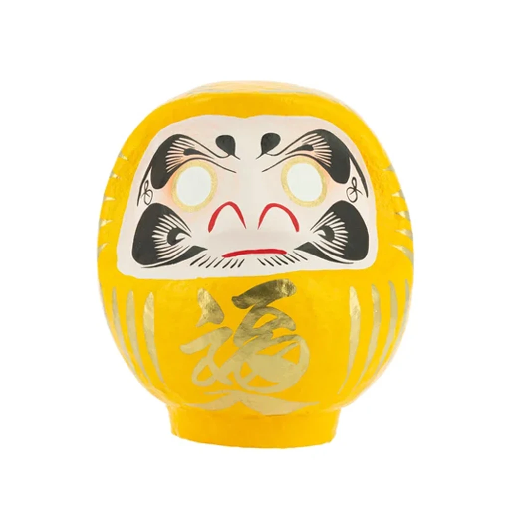 Daruma tradicional amarillo con rostro blanco, cejas y bigote negros, ojos sin pupilas y un carácter dorado en el frontal. Daruma tradicional amarillo con rostro blanco, cejas y bigote negros, ojos sin pupilas y un carácter dorado en el frontal.