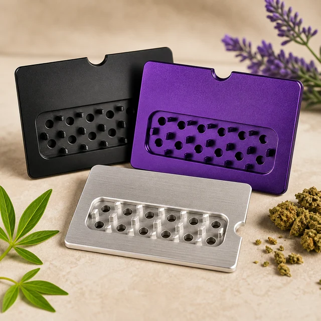 Tres grinders de tarjeta en negro, morado y plateado, con perforaciones y dientes, junto a hojas verdes y cogollos. Tres grinders de tarjeta en negro, morado y plateado, con perforaciones y dientes, junto a hojas verdes y cogollos.