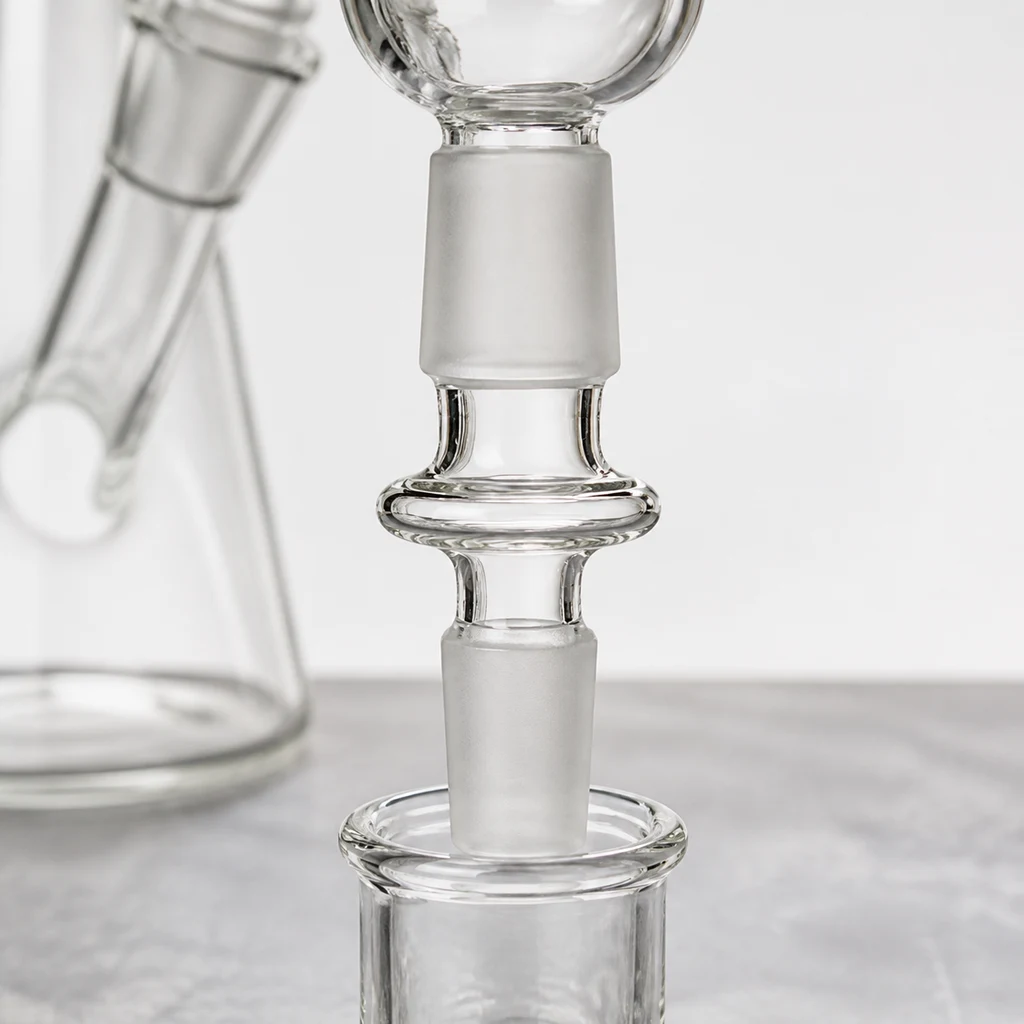 Primer plano de un adaptador transparente para bong, con extremos esmerilados y un aro central, colocado entre dos piezas de cristal.