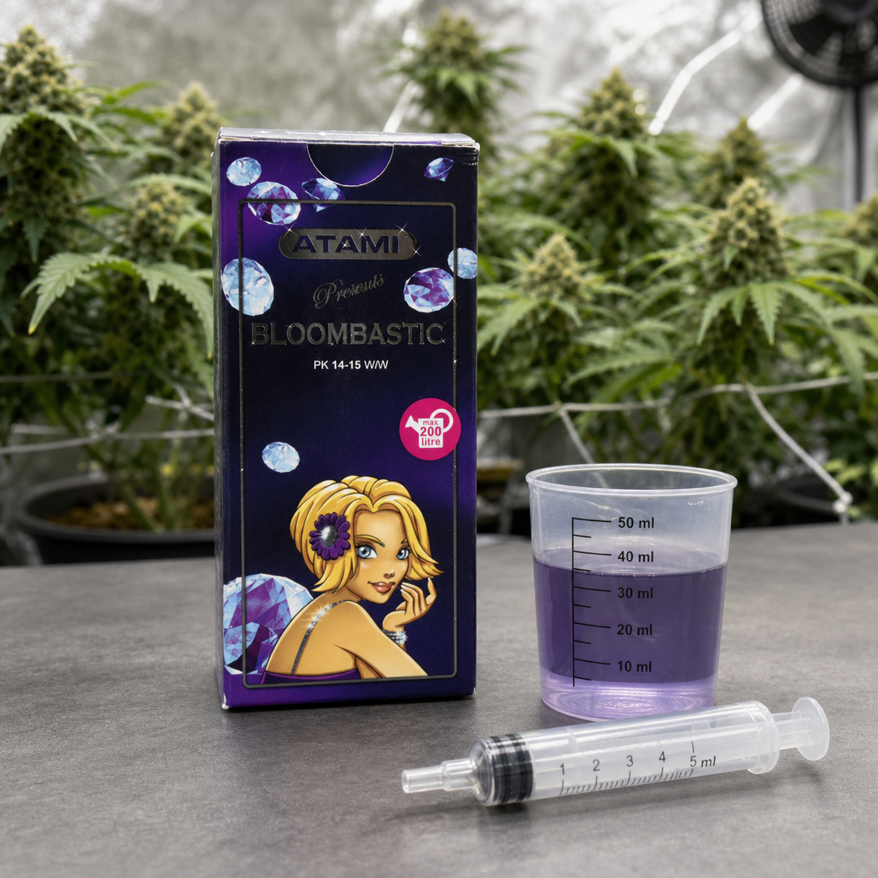 Caja de ATAMI Bloombastic junto a un vaso graduado con líquido violeta y una jeringa, con plantas de cannabis al fondo. Caja de ATAMI Bloombastic junto a un vaso graduado con líquido violeta y una jeringa, con plantas de cannabis al fondo.