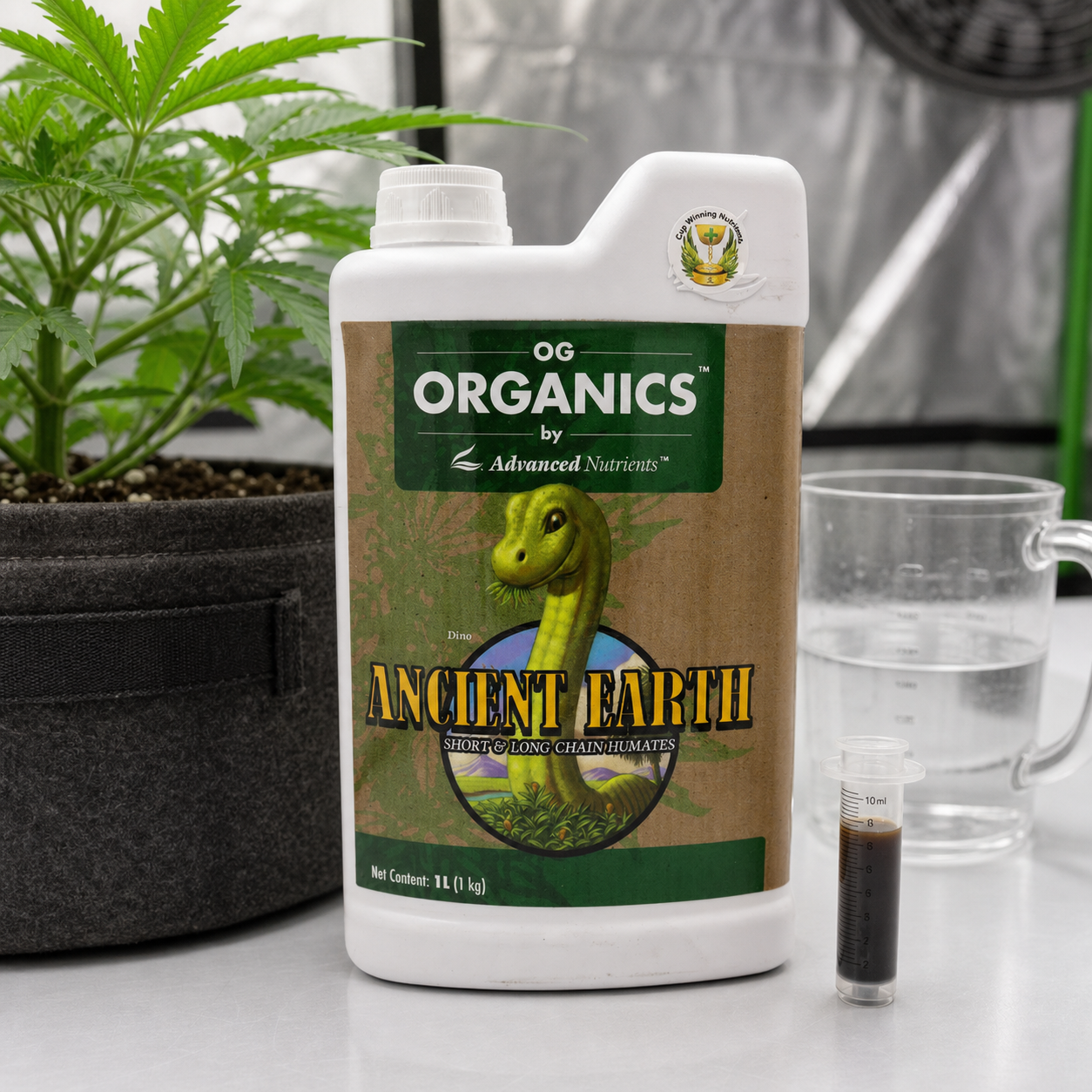 Envase de OG Organics Ancient Earth de Advanced Nutrients junto a una planta en maceta, una jarra con agua y una jeringa con líquido oscuro. Envase de OG Organics Ancient Earth de Advanced Nutrients junto a una planta en maceta, una jarra con agua y una jeringa con líquido oscuro.