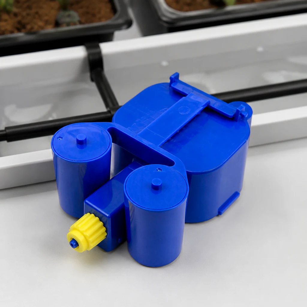 AquaValve AutoPot azul con cuerpos cilíndricos y conector amarillo, sobre una bandeja blanca junto a tubos negros y macetas.