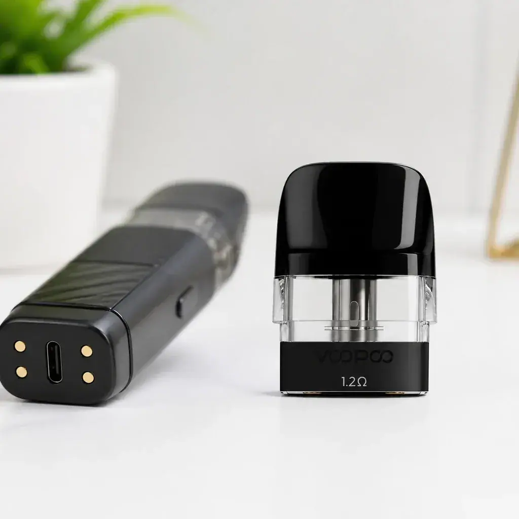 Cartucho Voopoo con boquilla negra, depósito transparente e inscripción de 1,2 Ω, junto a un dispositivo tumbado con puerto USB-C visible.