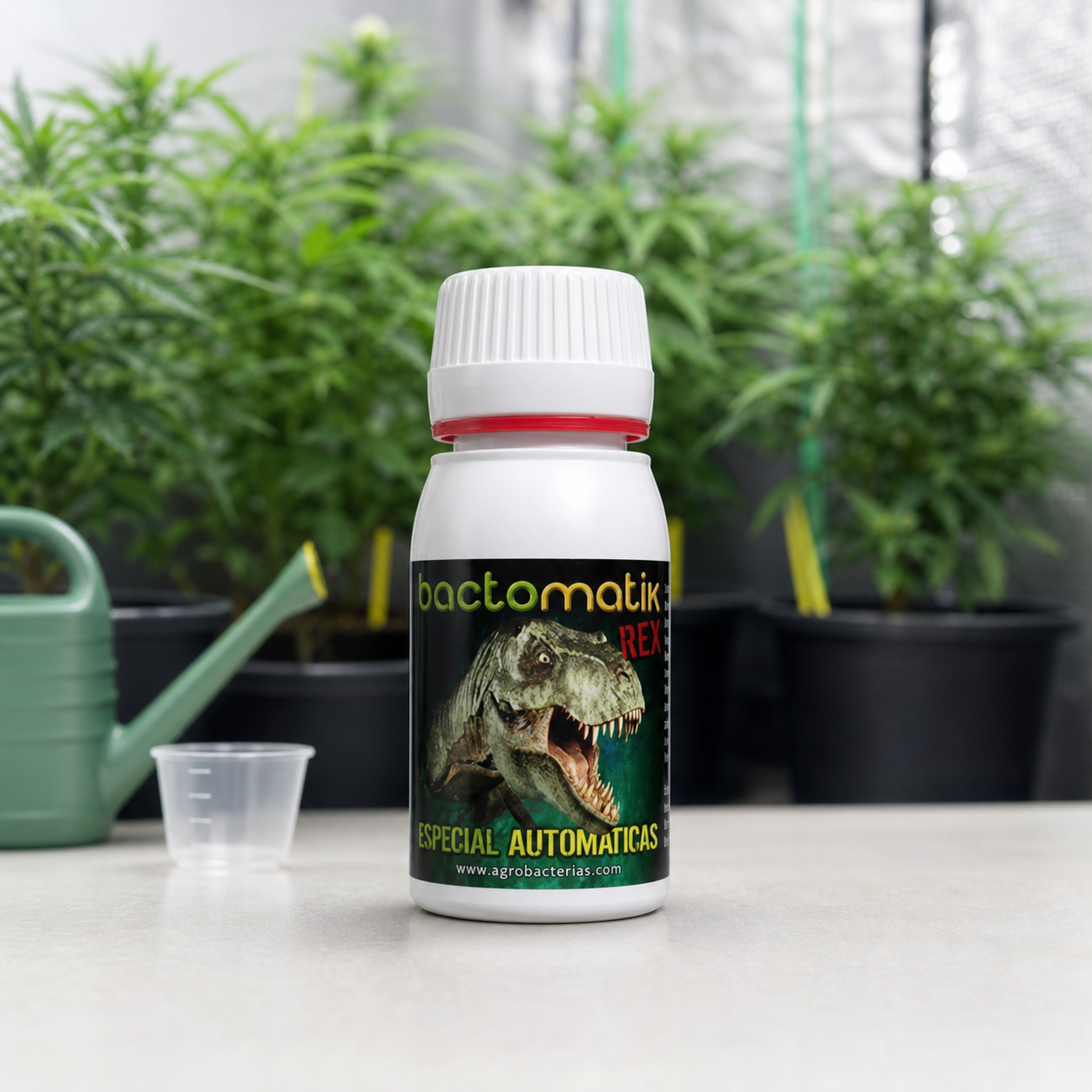Bote blanco Bactomatik Rex con un dinosaurio en la etiqueta, junto a un vaso dosificador y una regadera, con plantas de fondo.