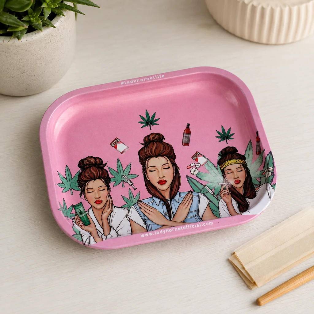 Bandeja de liar rosa ilustrada con mujeres y hojas de cannabis, sobre una mesa clara junto a papeles de liar.