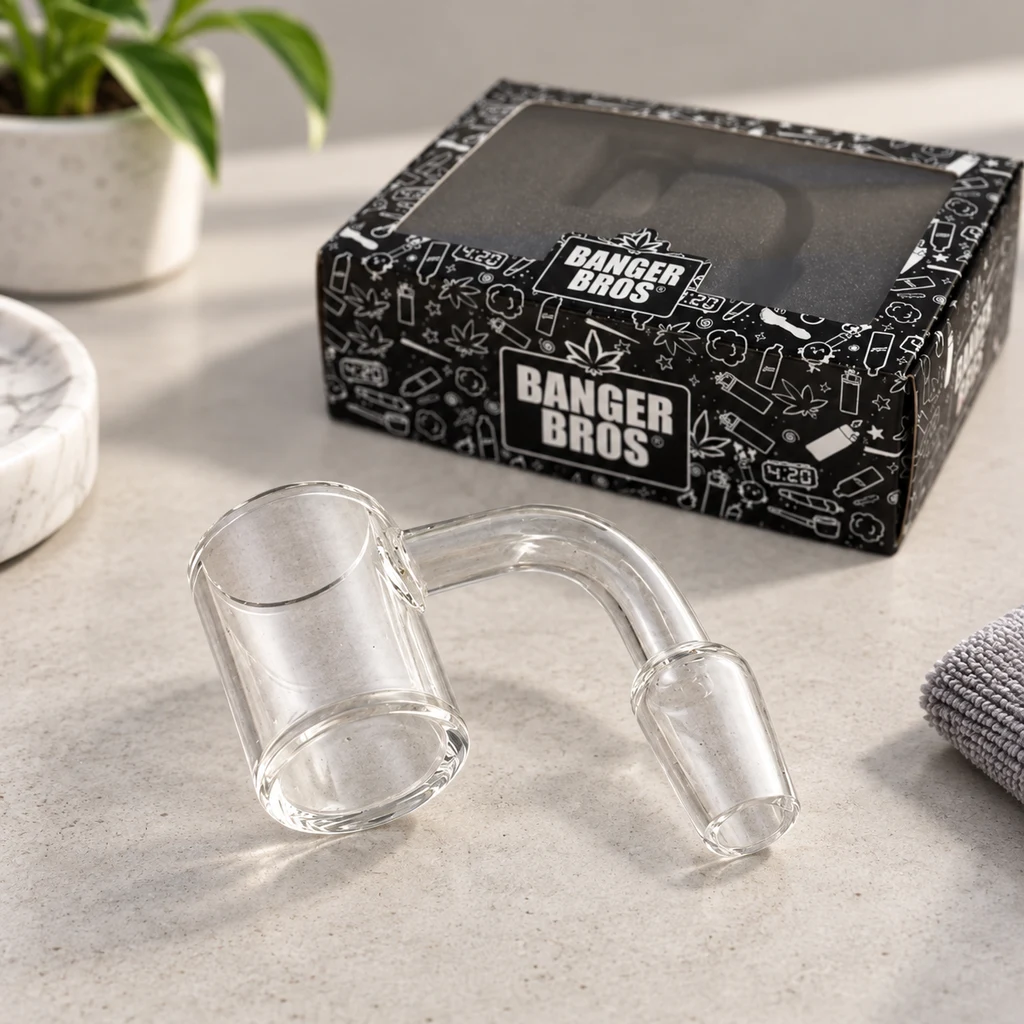 Banger transparente con recipiente cilíndrico, tubo curvado y conexión cónica sobre una mesa, junto a una caja negra Banger Bros.