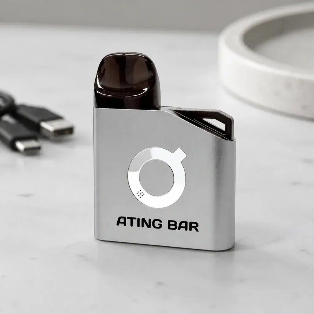 Pod Ating EVO-1 gris con boquilla oscura, logotipo circular y texto «ATING BAR», sobre una mesa con conectores USB al fondo.