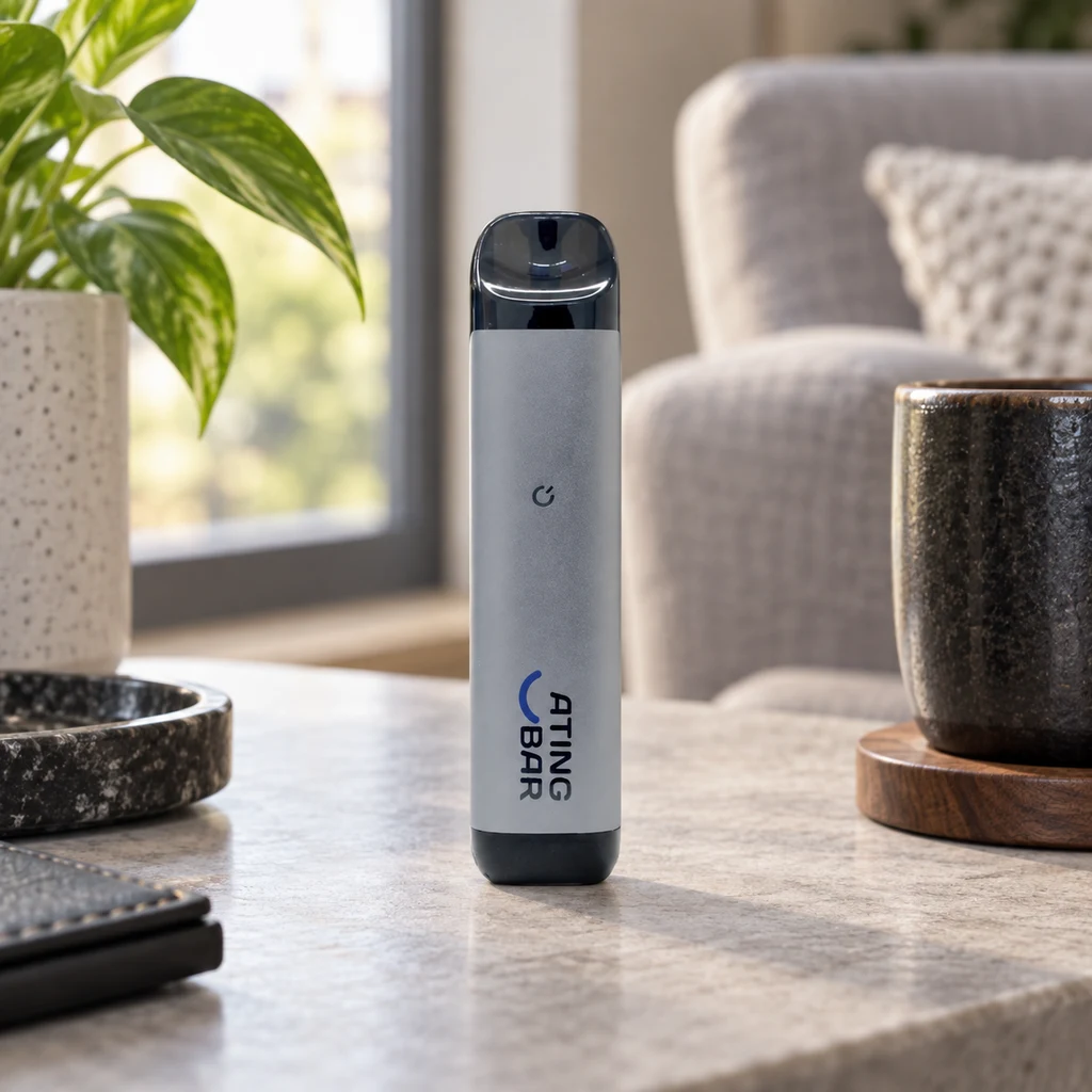 Batería de vapeo Ating EVO-2 gris, de pie sobre una mesa, con extremo superior oscuro y logotipo frontal, junto a una taza y una planta.
