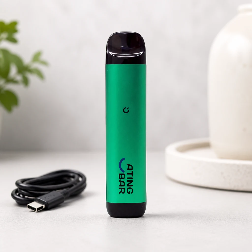 Dispositivo de vapeo verde con extremos negros y texto Ating Bar, colocado de pie junto a un cable USB negro enrollado.