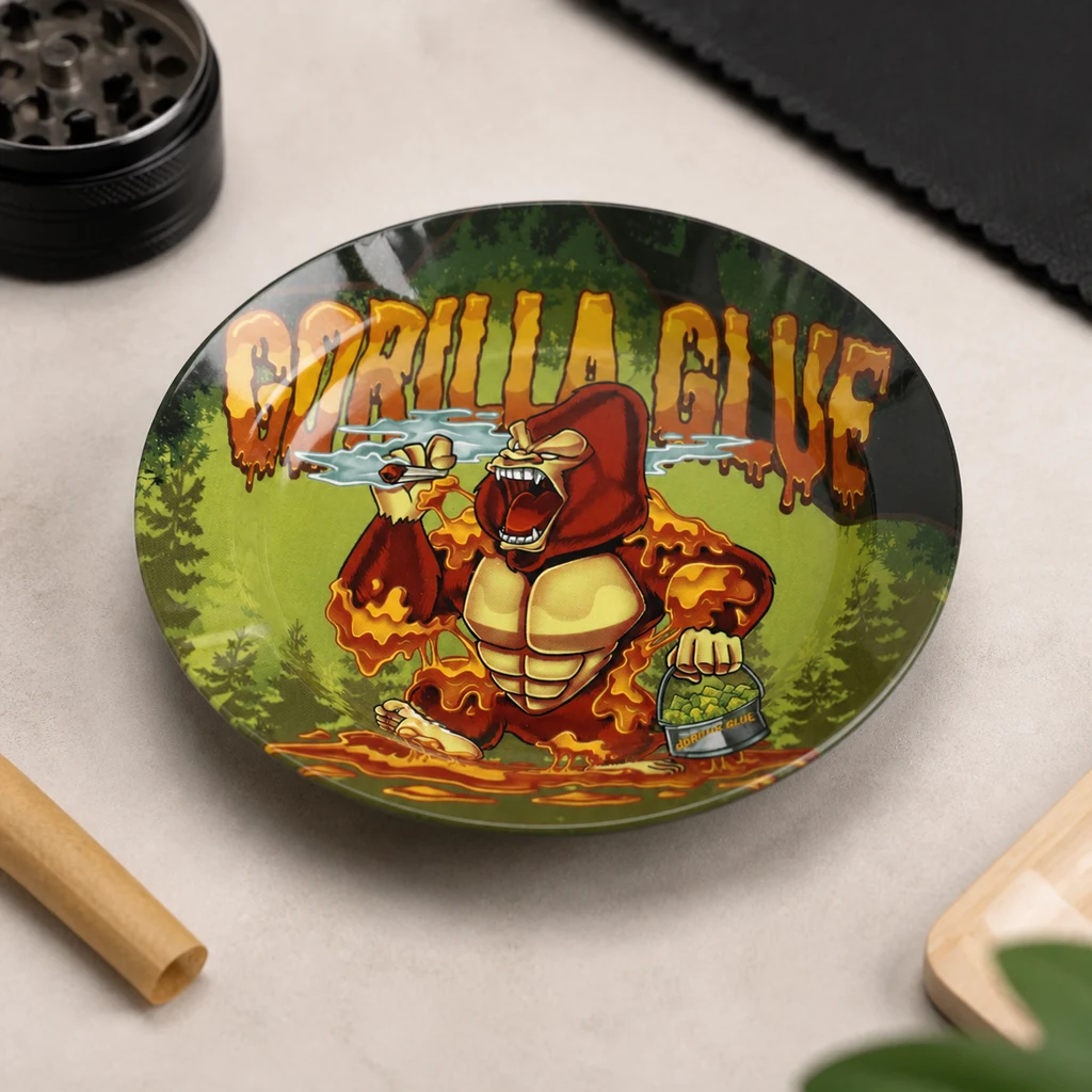 Cenicero redondo con la inscripción Gorilla Glue y un gorila ilustrado sobre fondo verde, junto a un grinder y papel enrollado.
