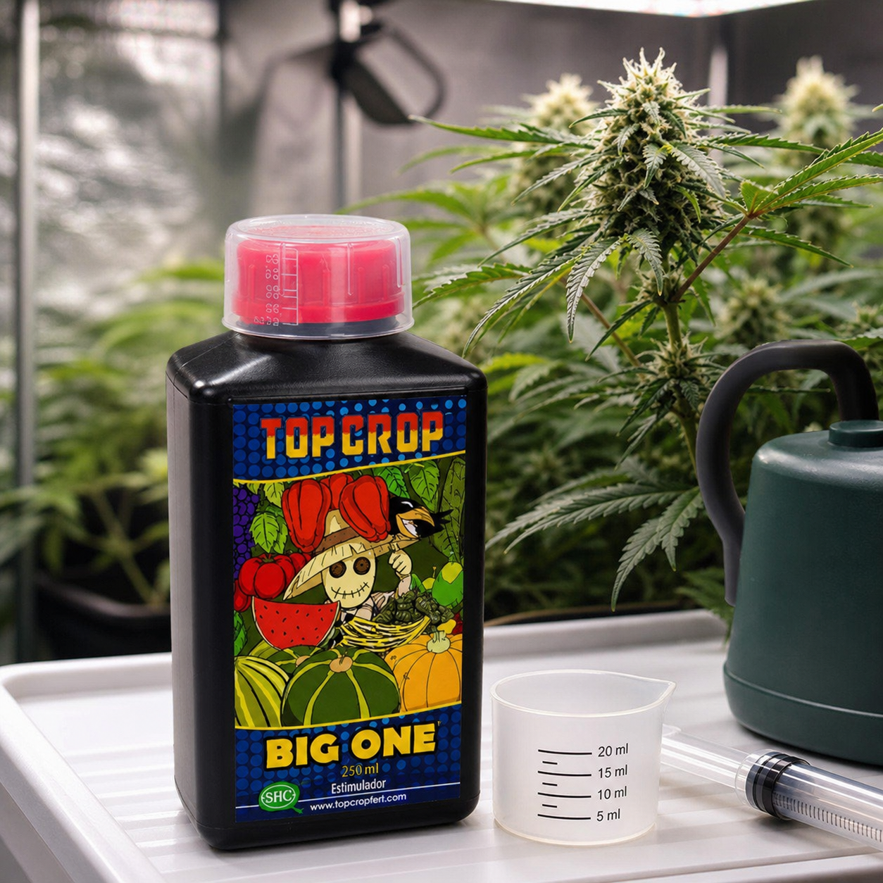Botella de Big One Top Crop con tapón rojo, junto a un vaso dosificador y una jeringa, con regadera y plantas en floración al fondo.