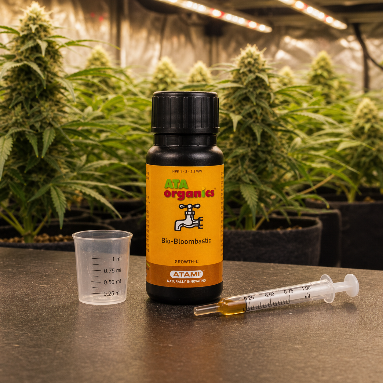 Bote de Bio-Bloombastic de Atami con etiqueta amarilla, junto a un vaso medidor y una jeringa, con plantas de cannabis al fondo. Bote de Bio-Bloombastic de Atami con etiqueta amarilla, junto a un vaso medidor y una jeringa, con plantas de cannabis al fondo.