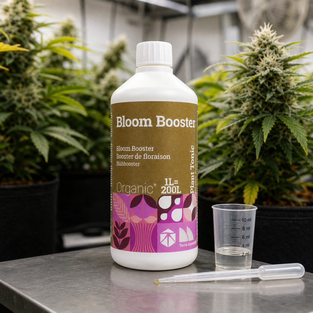 Botella de Bloom Booster con etiqueta marrón y rosa, junto a un vaso graduado y una pipeta, con plantas en floración al fondo.