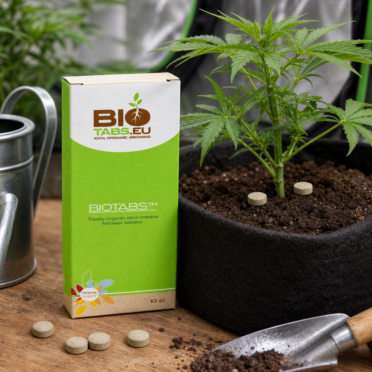 Caja de BioTabs junto a una planta de cannabis en maceta textil, con tabletas sobre la tierra y la mesa, una regadera y una pala.
