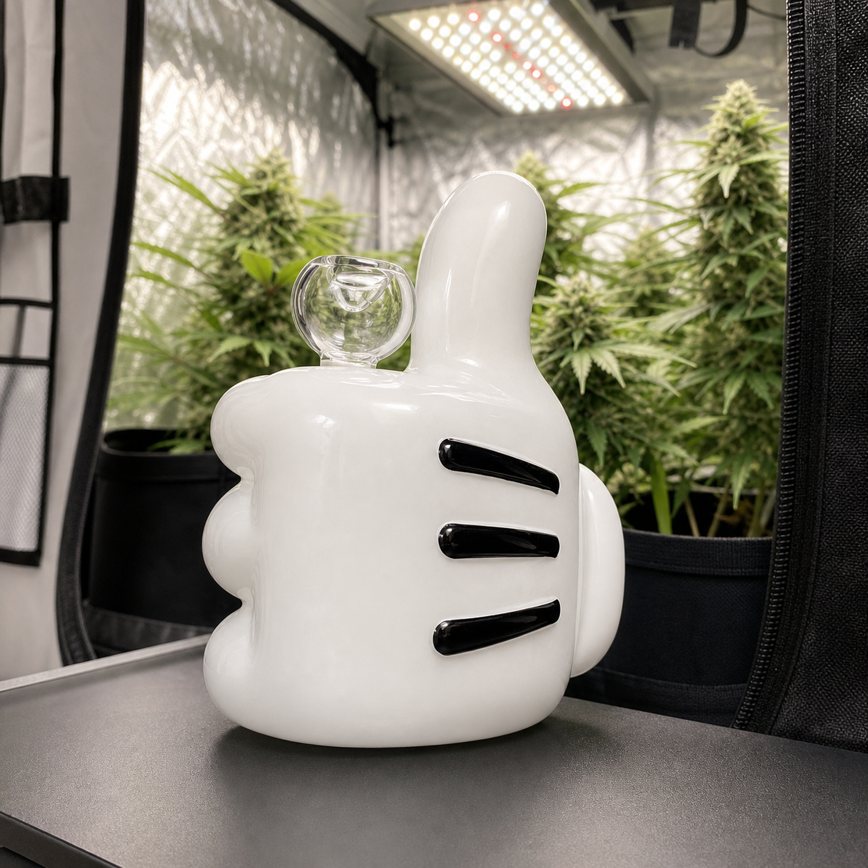 Bong Cartoon High Jinx blanco con forma de mano con pulgar levantado, detalles negros y cazoleta transparente, ante un armario con plantas.