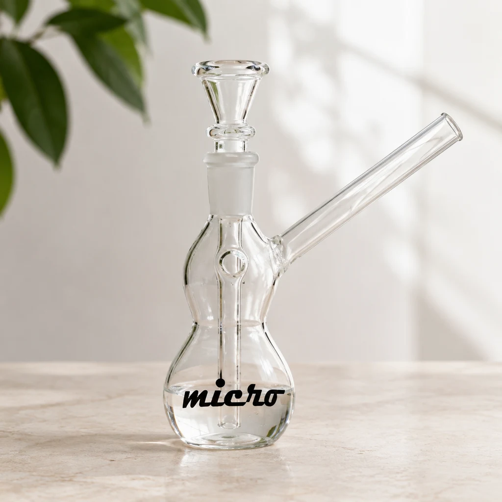 Bong de cristal transparente Micro con cuerpo curvado, cazoleta superior y boquilla lateral inclinada, sobre una mesa clara.